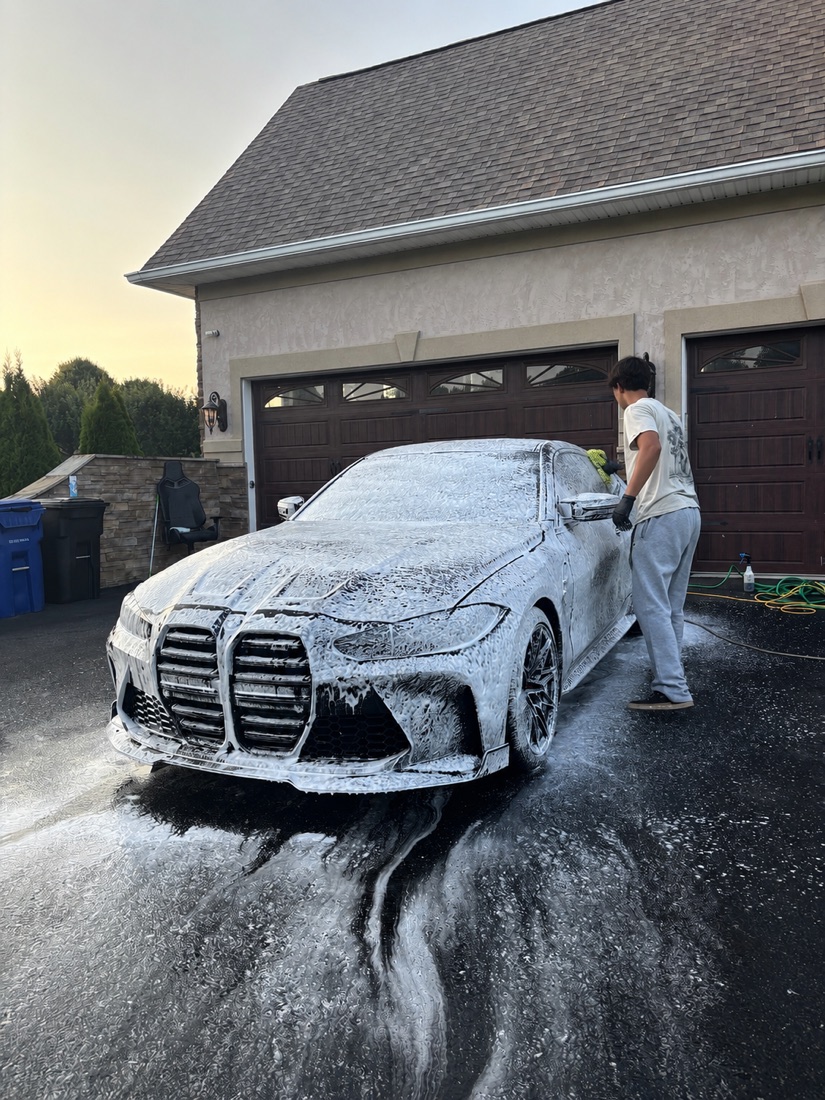 BMW M4 covered in foam being hand washed in a driveway at sunset.